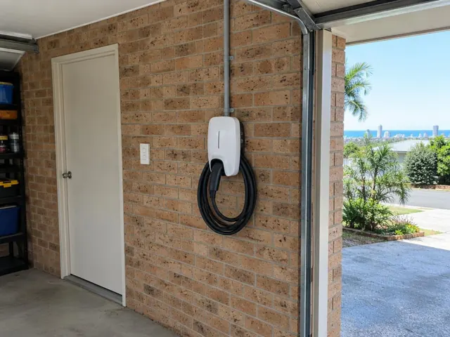 Wall-mounted EV charger with coiled cable on a brick garage wall beside a door in Burleigh Heads