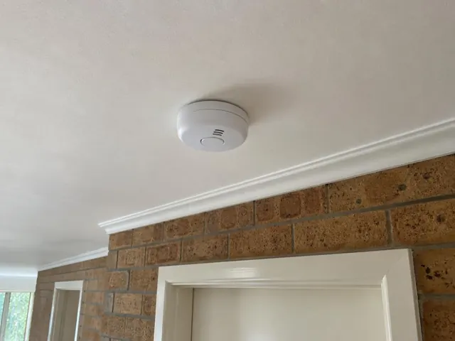 White smoke alarm on a white ceiling above a brick wall and doorway in Currumbin Waters